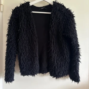 Svart pälsjacka från Gina Tricot - Säljer en svart pälsjacka från Gina Tricot med lång luddig fuskpäls och öppen front. Perfekt för dig som vill ha något extra till din outfit.