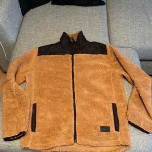 Brun teddyjacka från True North XL - Mysig brun teddyjacka från True North i storlek XL. Jackan har svarta detaljer på axlar och fickor, hel dragkedja framtill och hög krage. Perfekt för kyliga dagar när du vill vara både varm och snygg.