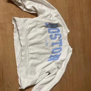 Vit sweatshirt Boston Kappahl - Vit sweatshirt från Kappahl med ljusblå text 'BOSTON' framtill. Tröjan har rund hals, långa ärmar och breda muddar vid ärmslut och nederkant. Mjuk insida och avslappnad passform, perfekt för en chill look.