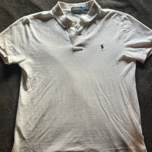 Vit slim fit piké från Ralph Lauren - Vit kortärmad pikétröja från Polo Ralph Lauren i slim fit-modell. Klassisk krage med två knappar och broderad logga på bröstet. Tillverkad i bomull för en skön och luftig känsla. Perfekt till jeans eller shorts för en clean och stilren look.