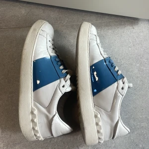 Valentino Garavani Open sneakers vit/blå - Snygga Valentino Garavani Open sneakers i vitt skinn med en bred blå lackad rem över mitten. Klassisk rund tå, vita snören och ikoniska nitar på hälen. Perfekta för dig som vill ha en stilren men ändå unik look.