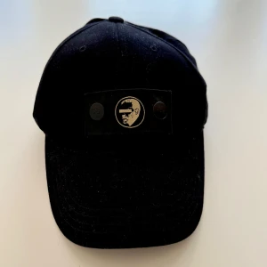 Svart keps med broderad logga - Svart keps i klassisk modell med böjd skärm och justerbar snapback baktill. Framtill sitter en broderad logga i vitt och svart på en patch, samt två svarta knappar. Liten detalj med röd, vit och blå rand bak. Perfekt för en cool och avslappnad stil.