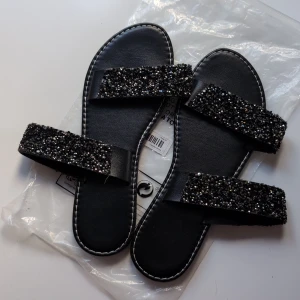 Svarta sandaler med glittriga band - Svarta sandaler med två breda band täckta av svarta och silvriga glittrande stenar. Platt sula och vita sömmar längs kanten ger en snygg kontrast. Perfekta för att lyfta vilken outfit som helst med extra bling.