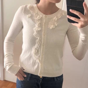 Vit kofta med blommiga detaljer Hollister - Supersöt vit kofta från Hollister med blommiga spetsdetaljer längs knappraden. Långärmad och i mjukt, tunt material som känns skönt mot huden. Perfekt till jeans eller kjol för en romantisk look. Klassisk passform och rund halsringning.