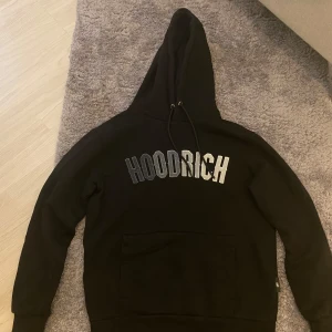 Svart hoodie från Hoodrich med tryck - Svart hoodie från Hoodrich i storlek S med stor logga broderad i vitt och grått över bröstet. Klassisk känguruficka framtill, snörning i huvan och ribbade muddar. Perfekt för en streetwear-look.