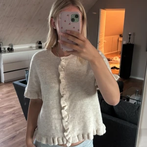 Beige stickad kofta med volang - Supersöt beige stickad kofta med kort ärm och volangkant längs både knäppning och nederkant. Snygga pärlknappar framtill och mjukt material som känns riktigt cozy. Perfekt att slänga på sig över ett linne eller t-shirt för en gullig look.