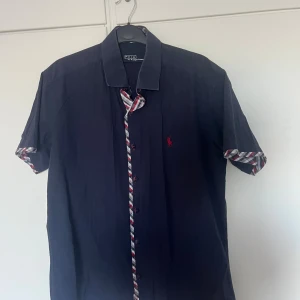 Mörkblå kortärmad skjorta Polo Ralph Lauren - Snygg mörkblå skjorta från Polo Ralph Lauren med korta ärmar och rutiga detaljer i rött, vitt och blått vid knappslå och ärmslut. Klassisk krage och röd broderad logga på bröstet. Perfekt för dig som vill ha en stilren men ändå unik look.
