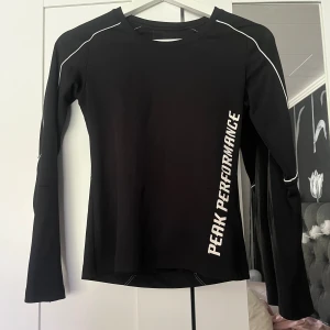 Svart långärmad träningströja Peak Performance - Svart långärmad träningströja från Peak Performance med vit logga längs sidan och vita detaljer på axlarna. Tight passform och tillverkad i ett stretchigt syntetmaterial som andas, perfekt för träning eller sportiga aktiviteter.