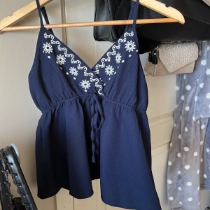 Mörkblå broderad linnetopp - Supersöt mörkblå linnetopp  med vita broderade blommor längs v-ringningen. Toppen har smala axelband, knytning framtill och en lätt utställd passform🤍💙