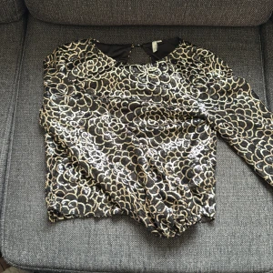 Paljettblus i svart och guld från Nelly storlek 34 - Glittrig långärmad blus från Nelly i svart tyg täckt av guld- och silverpaljetter i blommigt mönster. Blusen har rund halsringning och en knappdetalj i nacken. Fint skick!