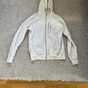 Vit hoodie från Sail Racing med dragkedja - Vit hoodie från Sail Racing i storlek M. Tröjan har hel dragkedja framtill, huva med snörning och en bröstficka med zip. Mjuk insida i fleece och ribbade muddar vid ärmslut och nederkant. Perfekt för chill eller när du vill vara extra bekväm.