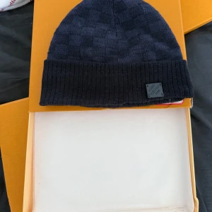 Mörkblå Louis Vuitton mössa - Snygg mörkblå mössa från Louis Vuitton med diskret rutigt mönster och ribbad kant. Märkets logga sitter på en liten patch framtill. Perfekt accessoar för kyliga dagar och en stilren look. Använd 1 gång. Vid frågor kontakta mig. 