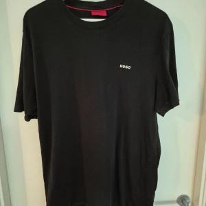 Svart t-shirt från Hugo - En stilren svart t-shirt från Hugo med diskret logga på bröstet. Klassisk rund halsringning och korta ärmar. Tillverkad i mjuk bomull som känns skön mot huden. Perfekt för en clean och enkel look.