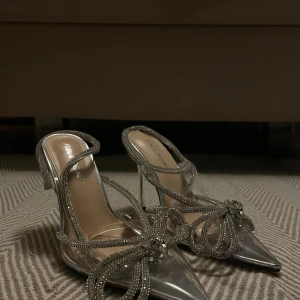 Glittriga silverklackar från Public Desire - Säljer ett par riktigt snygga klackar från Public Desire i silver med spetsig tå och höga smala klackar. Skorna har glittrande remmar dekorerade med strass och en stor rosett framtill. Perfekta för att sticka ut och ge extra bling till din outfit.