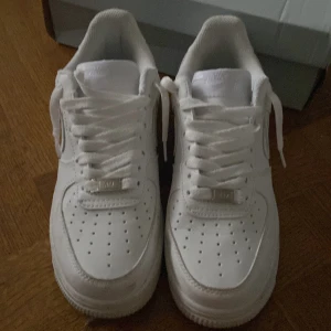 Nike Air Force 1 helvita sneakers - Klassiska Nike Air Force 1 sneakers i helvitt skinn med perforerad tå, rund tå och platt sula. Skorna har snörning och den ikoniska AF1-detaljen på snörena. Perfekta för dig som vill ha en clean och tidlös look.