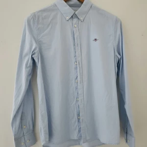 Ljusblå skjorta från Gant - Snygg oanvänd ljusblå skjorta från Gant med klassisk button-down krage och broderad logga på bröstet. Skjortan har långa ärmar, vita knappar och är gjord i mjuk bomull. Perfekt för dig som gillar stilrena och tidlösa plagg. Storlek 158/164.