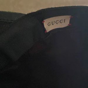 Svart keps från Gucci med GG-mönster - Snygg svart keps från Gucci i storlek M med det klassiska GG-mönstret över hela kepsen. Kepsen har en rundad form och är tillverkad i ett slitstarkt syntetmaterial med detaljer i svart. Perfekt för dig som vill ha en lyxig och trendig accessoar.