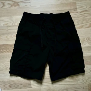 Svarta cargo shorts från H&M - Svarta cargo shorts från H&M med regular fit och snörning i midjan. Shortsen har praktiska sidofickor med lock och är gjorda i lätt polyester, perfekt för chill dagar. Enkel och clean design som funkar till sneakers eller slides.