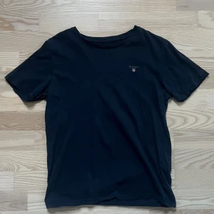 Svart t-shirt från GANT i bomull - Svart t-shirt från GANT med klassisk rund hals och liten broderad logga på bröstet. Tillverkad i mjuk bomull som känns skön mot huden. Passformen är normal och ärmarna är korta, perfekt för en chill och stilren look. Ett litet hål som är på bilden . 