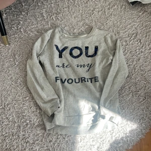 Grå sweatshirt med text och paljetter - Mysig grå sweatshirt med texten 'YOU are my FAVOURITE' framtill. Ordet 'YOU' är dekorerat med mörkblå paljetter som ger en snygg detalj. Tröjan har rund halsringning och långa ärmar, perfekt för chill dagar.