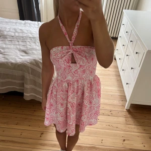 Rosa och vit kortklänning med knyt - Supersöt kort klänning i rosa och vitt med blommigt mönster. Klänningen har en omlottknytning vid bysten som kan bäras både som halterneck eller axelbandslös. Utsvängd kjol och lätt material, perfekt för varma dagar.