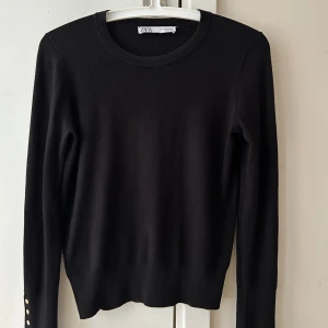 Svart stickad tröja från Zara - En svart stickad tröja från Zara med rund halsringning och långa ärmar. Tröjan har snygga guldfärgade knappar vid ärmsluten som extra detalj. Perfekt att styla med jeans eller kjol för en clean look.