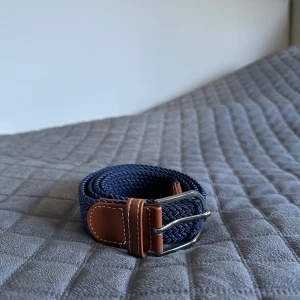 Mörkblå flätat bälte med skinn - Snyggt mörkblått flätat bälte med bruna skinn-detaljer och silverspänne. Bältet har en stilren look med vit kontrastsöm på skinnet och passar perfekt till jeans eller chinos. Materialet är mjukt och flexibelt, vilket gör det bekvämt att bära hela dagen.