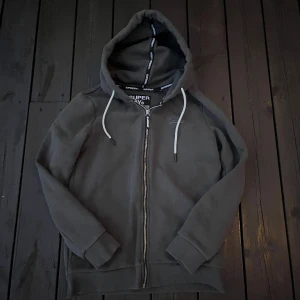 Mörkgrå hoodie från Superdry med dragkedja - Säljer en mörkgrå hoodie från Superdry i storlek 34. Tröjan har huva med vita snören, dragkedja framtill och diskret Superdry-logga på bröstet. Perfekt för chill dagar och enkel att matcha med jeans eller leggings.