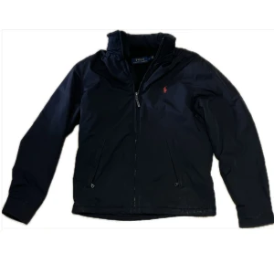 Svart Ralph Lauren jacka - Snygg svart vindjacka från Polo Ralph Lauren med klassisk röd logga på bröstet. Jackan har dragkedja framtill, ståkrage och två fickor med dragkedja. Perfekt för blåsiga dagar och enkel att matcha med allt.