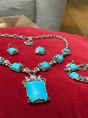 Turkos och silverfärgad väst - Säljer en unik väst med rektangulära turkosa detaljer och silverfärgade metallinslag. Västen har ett coolt, bohemiskt uttryck med dekorativa mönster och kedjor. Perfekt för dig som vill sticka ut och addera färg till din outfit.