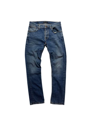 Blå jeans från Nudie Jeans Co, 31/32 - Säljer ett par klassiska blå jeans från Nudie Jeans Co i storlek 31/32. Jeansen har fem fickor, orangea kontrastsömmar och en rak passform. Materialet är slitstarkt denim i bomull och färgen är snyggt tvättad blå med ljusare partier. Perfekt för dig som gillar tidlös stil.