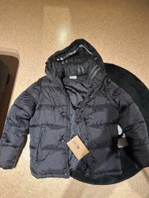 Svart pufferjacka från Burberry - Säljer en svart pufferjacka från Burberry med huva och snygga detaljer som märkets logga på bröstet och knappar med Burberry-tryck. Jackan har dragkedja och knappar framtill, samt fickor med dragkedja. Insidan har ett grått rutigt foder som ger extra stil.