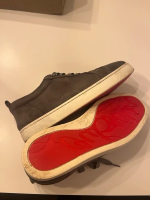 Gråa sneakers från Christian Louboutin - Säljer ett par gråa sneakers från Christian Louboutin med klassisk röd sula och vit kant. Skorna har snörning och är tillverkade i mocka med rund tå. Perfekt för dig som gillar lyxiga detaljer och vill sticka ut med den ikoniska röda sulan.