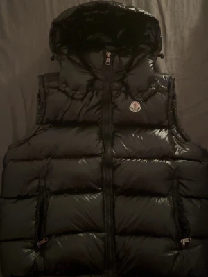 Svart dunväst från Moncler med huva - Säljer en svart, glansig dunväst från Moncler med huva och två dragkedjefickor. Västen har quiltad design, Moncler-logga på bröstet och innerfoder med serietidningsmönster. Perfekt för dig som vill ha en snygg och varm väst med streetkänsla.