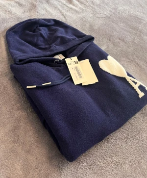 Ami Paris tröja  - Snygg marinblå hoodie från Ami paris med vit hjärtdetalj och bokstaven A på bröstet. Tröjan har huva med snörning, ribbade muddar och en praktisk magficka. Perfekt för dig som gillar stilrena och coola plagg.