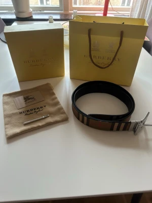 Rutig Burberry bälte med silverdetaljer - Säljer ett snyggt Burberry bälte med klassiskt rutigt mönster i beige, brun, svart och rosa. Bältet har en rejäl silverfärgad spänne med Burberry-logga. Kommer med originalpåse, certifikat och box. Perfekt för dig som vill ha en ikonisk accessoar.