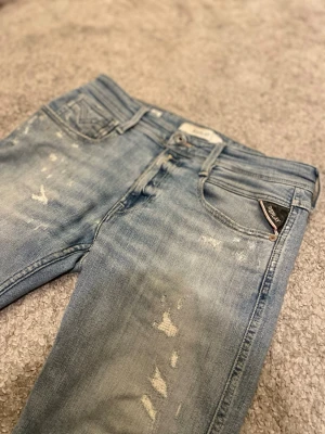 Replay anbass jeans med slitningar - Säljer ett par ljusblå Replay jeans med skön passform och snygga slitningar framtill. Klassisk femficksmodell med Replay-logga på fickan. Jeansen är tillverkade i mjukt denimtyg med stretch för extra komfort och har en modern, smal siluett.