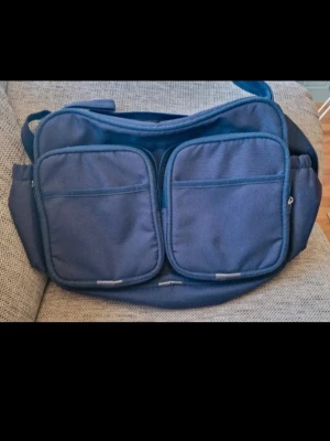 Blå skötväska - Säljer en blå axelväska i polyester med flera praktiska fack och dragkedjor. Väskan har två stora fickor med dragkedja framtill och på dessa fack utan dragkedja, sidofack, ett rymligt huvudfack och ett stort fack med drakedja på baksida där det är en liten fläck (se bild). Inuti själva väskan finns det tre fack utan dragkedja och en plastficka.  Slitage syns på kanterna (se bild).