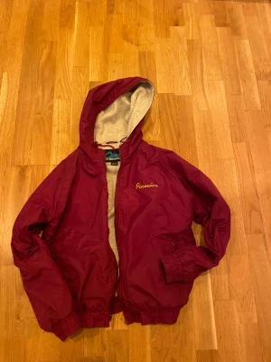 Burgundy vindjacka från Tri-Mountain - Säljer en burgundy vindjacka med huva från Tri-Mountain. Jackan har gul broderad text både fram och bak, med tryck 'Schaumburg Vikings 2001' och ett vikingmotiv på ryggen. Insidan av huvan är ljusgrå och jackan har dragkedja samt resår vid ärmslut och midja.