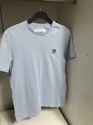 Ljusblå t-shirt från Lyle & Scott - Fräsch ljusblå t-shirt från Lyle & Scott med klassisk rund hals och korta ärmar. T-shirten har en gul broderad logga på bröstet och är tillverkad i 100% ekologisk bomull, vilket gör den både mjuk och skön. Perfekt för dig som gillar stilrena plagg med en clean vibe.