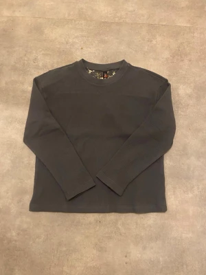 Mörkgrå långärmad tröja med broderi - Säljer en mörkgrå långärmad tröja med rund hals och ett snyggt broderat blommönster i vitt och beige på ryggen. Materialet är stickat och har en skön struktur som känns chill. Perfekt för dig som gillar unika detaljer och stilren design.