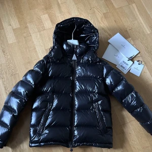 Mörkblå Moncler Maya - Säljer en svart, glansig pufferjacka från Moncler med huva och två fickor med dragkedja. Jackan har en klassisk Moncler-logga på ärmen och quiltad design. Perfekt för kalla dagar och har en snygg, modern look som sticker ut.