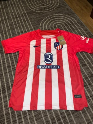 Rödvit randig Nike fotbollströja - Säljer en officiell Atlético Madrid fotbollströja från Nike med röda och vita vertikala ränder, klubbmärke på bröstet och sponsorloggor. Tröjan har korta ärmar och är tillverkad i lätt polyester som andas. Perfekt för dig som älskar fotboll och vill ha en snygg matchtröja.