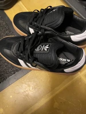 Adidas Samba sneakers svart/vit - Säljer ett par Adidas Samba sneakers i svart och vitt med klassiska tre ränder på sidan och gummisula. Skorna har snörning, rund tå och detaljer i skinn och mocka. Perfekt för dig som gillar retrostil och streetwear.