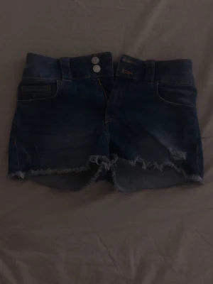 Blå jeansshorts med fransig kant - Säljer ett par blå jeansshorts med fransig och rå kant nertill. Shortsen har två knappar i midjan, klassiska fickor och en snygg slitning på benen. Perfekta för varma dagar och chill häng med kompisarna.