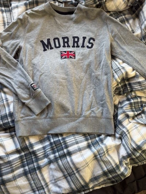 Grå sweatshirt från Morris XS - Snygg grå sweatshirt från Morris i storlek XS med broderad text och brittisk flagga på bröstet. Tröjan har rund hals och långa ärmar, perfekt för chill dagar. Mjuk bomullskänsla och klassisk stil med ribbade muddar vid ärmslut och nederkant.