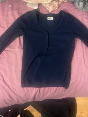 Mörkblå ribbad topp från Hollister - Säljer en mörkblå långärmad topp från Hollister i storlek S. Toppen har ribbad struktur, v-ringning och knappar framtill. Perfekt att styla med jeans eller kjol för en chill look.