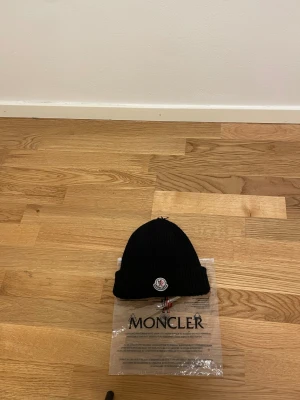 Svart Moncler mössa  - Säljer en svart ribbstickad mössa från Moncler med klassisk logotyp framtill. Mössan är tillverkad i mjuk ull och har en uppvikt kant för extra stilpoäng. Perfekt för dig som vill ha en clean och lyxig look under kyliga dagar.