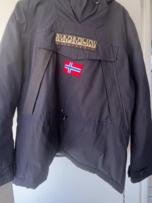 Svart jacka från Napapijri - Svart vindjacka från Napapijri med stor logga och norsk flagga på bröstet samt patch på ärmen. Jackan har huva, dragkedja i sidan och justerbar nederkant. Perfekt för dig som gillar streetwear och vill ha en snygg jacka med coola detaljer.        ‼️🚨PRISET ÄR INTE HUGGET I STEN🚨‼️