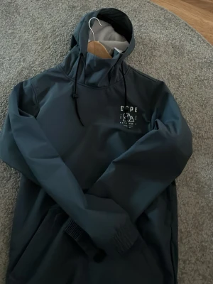 Mörkblå vindjacka från Dope - Snygg mörkblå vindjacka från Dope med stor vit logga och grafiskt tryck på både bröstet och ryggen. Jackan har huva med snörning och långa ärmar med elastiska muddar. Perfekt för blåsiga dagar och outdoor vibes.